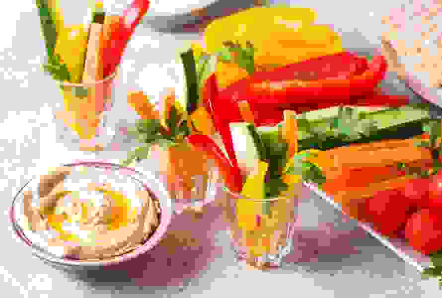 Un verre rempli de bâtonnets croquants de carottes, poivrons et courgettes, accompagné d’un bol de houmous crémeux. Le snack parfait pour une pause – riche en nutriments et prêt en quelques minutes

