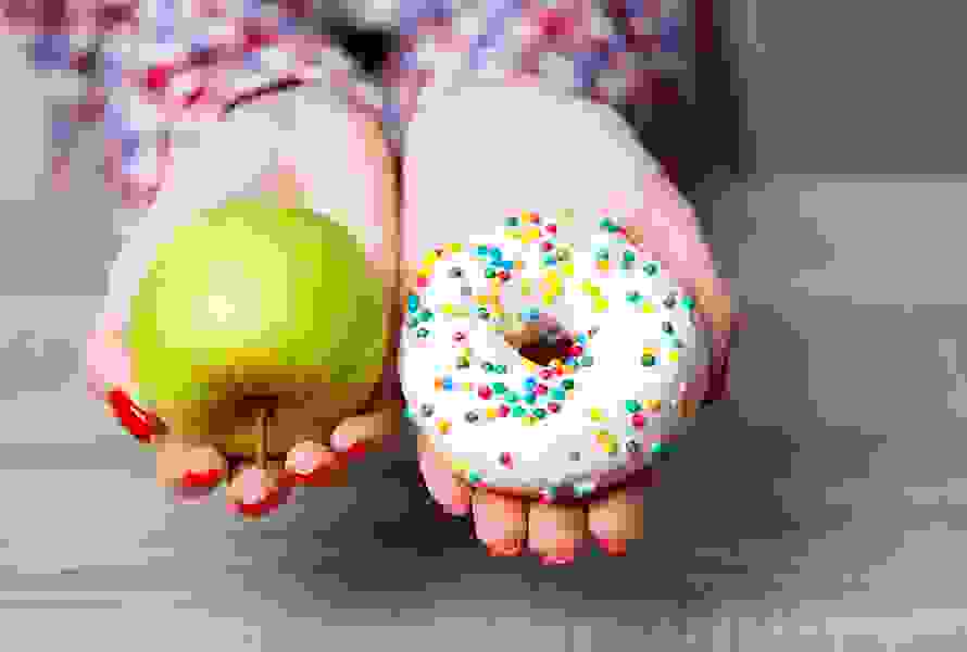 Eine Frau hält einen grünen Apfel in der einen und einen Donut mit weisser Glasur und bunter Deko in der anderen Hand – eine symbolische Darstellung der bewussten Ernährung in der Schwangerschaft.
