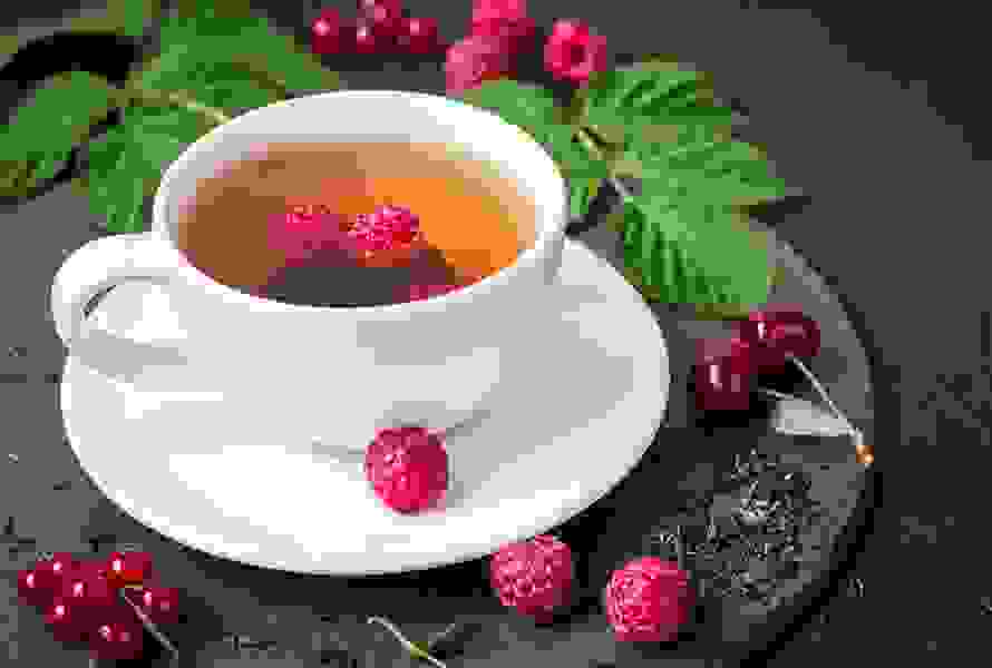 Eine dampfende Tasse Himbeerblättertee, dekorativ umgeben von frischen Himbeeren und Blättern – bitter-herb im Geschmack, aber hilfreich zur Geburtsvorbereitung.
