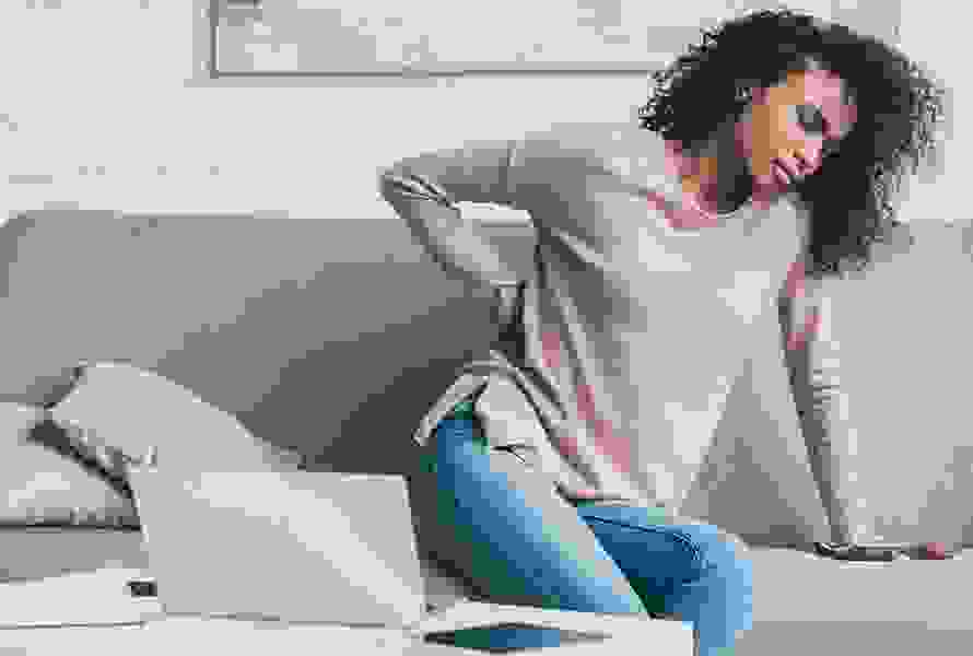 Eine Frau sitzt entspannt auf dem Sofa, hält ihren unteren Rücken – typisches Schwangerschaftssymptom und Tipp für mehr Wohlbefinden.