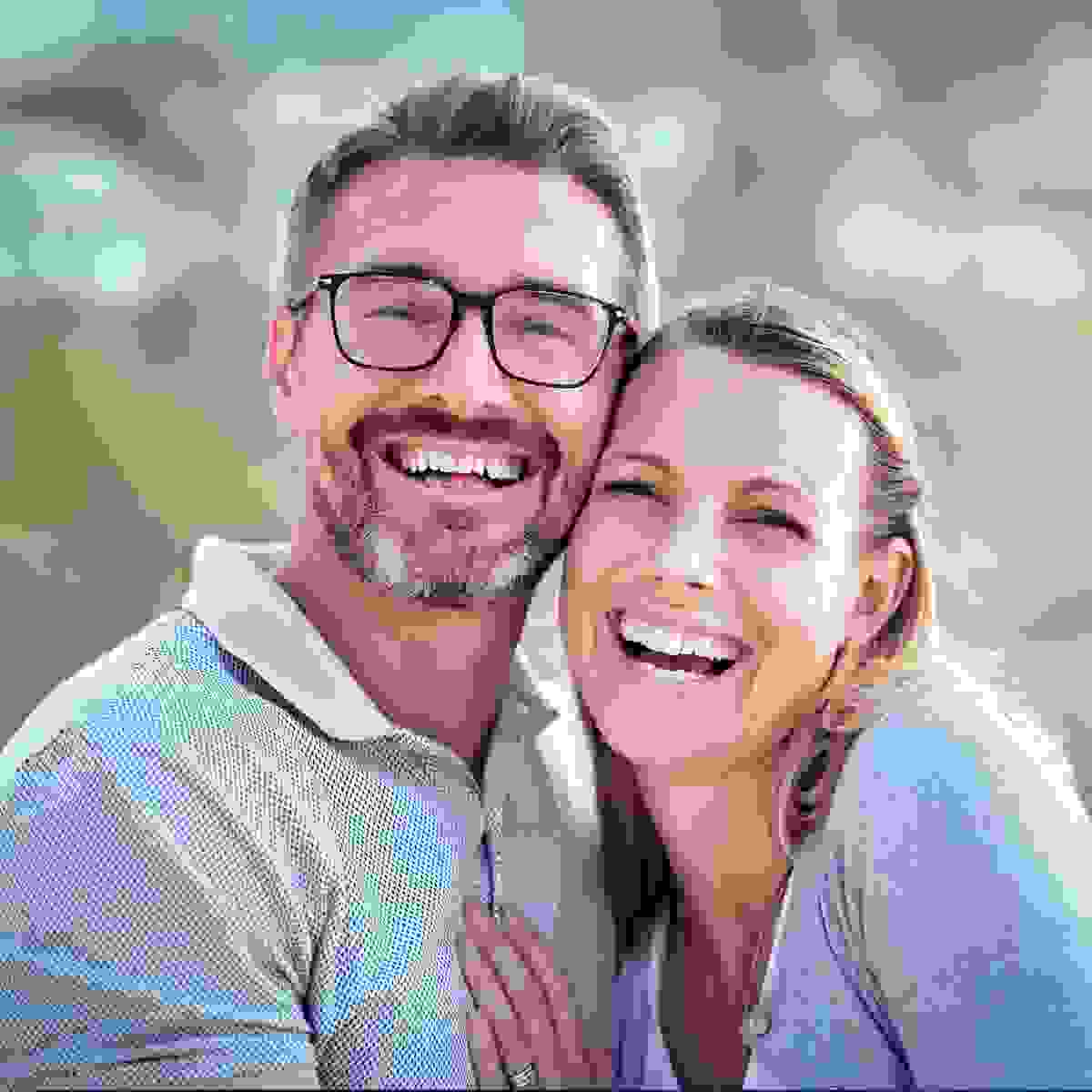 Zwei Personen stehen eng beieinander und lächeln in die Kamera. Die linke Person trägt eine Brille und ein helles Hemd, die rechte ein helles Oberteil. Der Hintergrund ist unscharf und zeigt eine Aussenkulisse mit Gebäuden und etwas Grün.