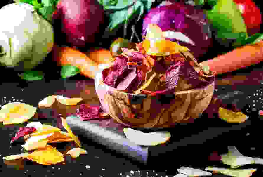 Un bol en bois rempli de chips de légumes maison colorés à base de betteraves, de tomates, de colraves et de carottes, placé devant des légumes frais sur une table en bois – pauvre en calories et croustillant.

