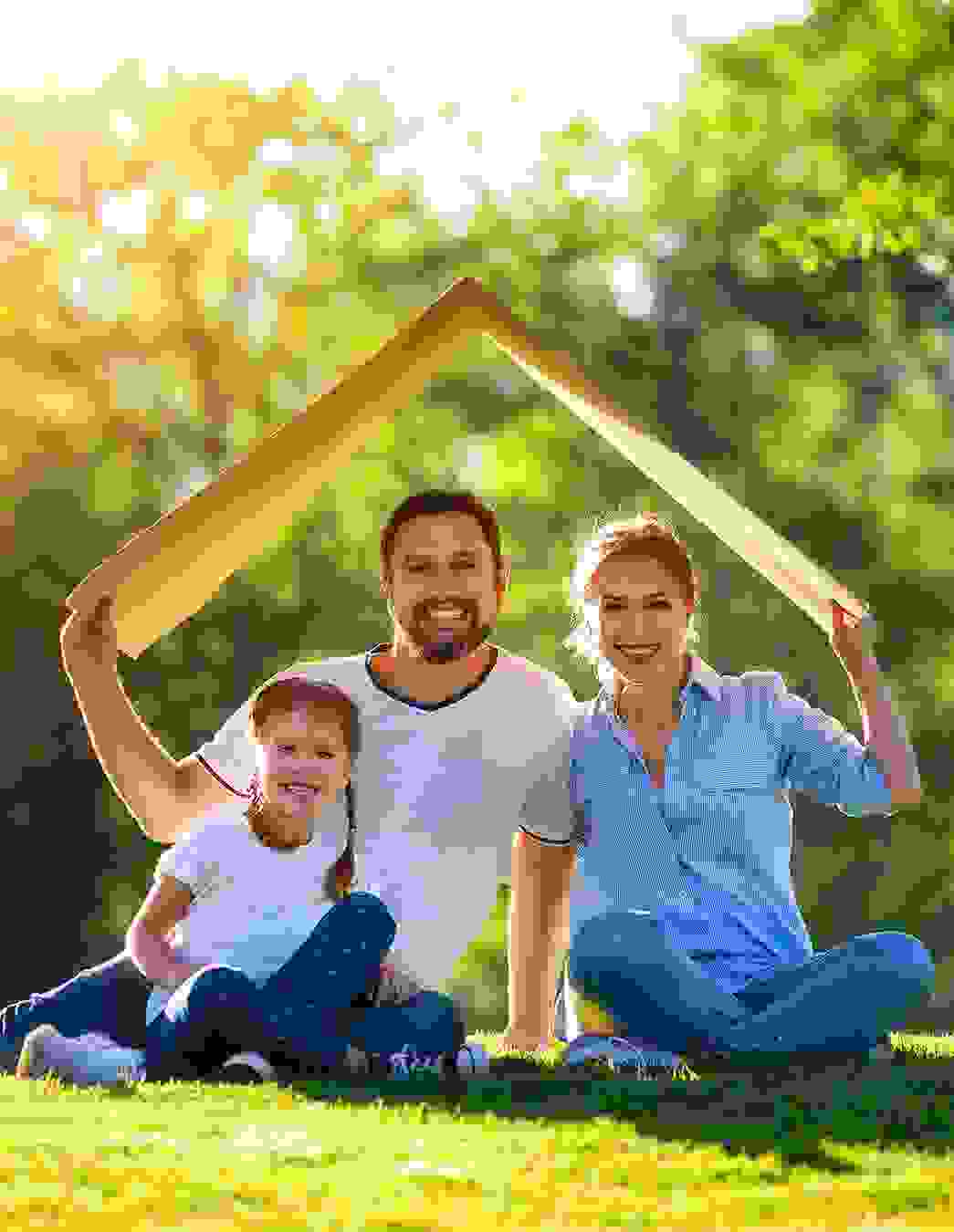 Ein Paar sitzt mit seiner kleinen Tochter auf einer grünen Wiese und hält zum Schutz ein Kartonstück in Form eines Hausdachs über ihre Köpfe – ein Symbol für Sicherheit und Geborgenheit.