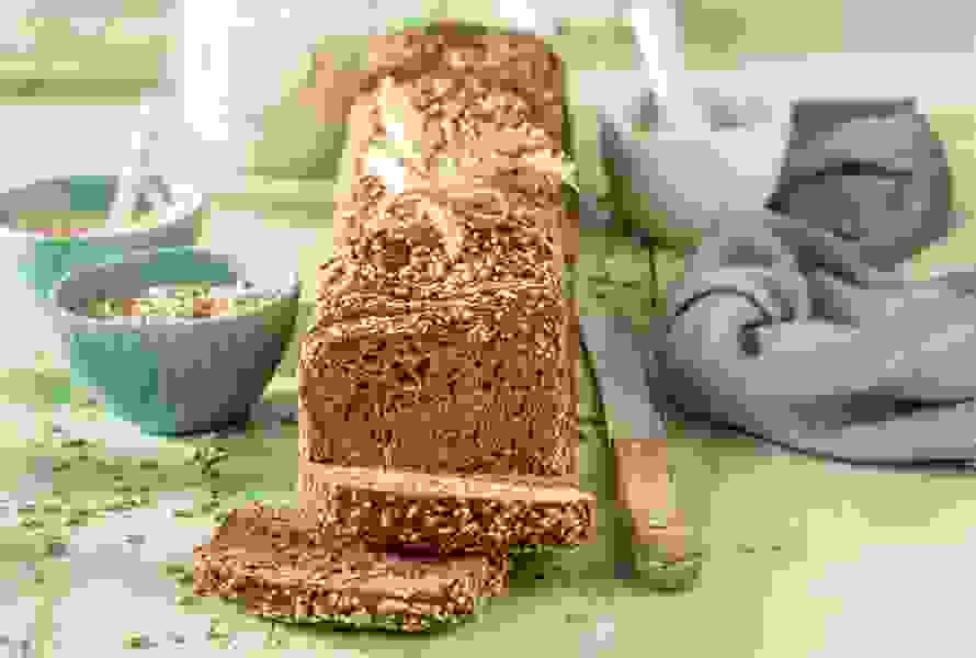 Kastenvollkornbrot mit zwei angeschnittenen Scheiben auf grünem Holztisch, daneben ein Brotmesser, Schüsseln mit Körnern und ein Glas – sättigend und gesund.