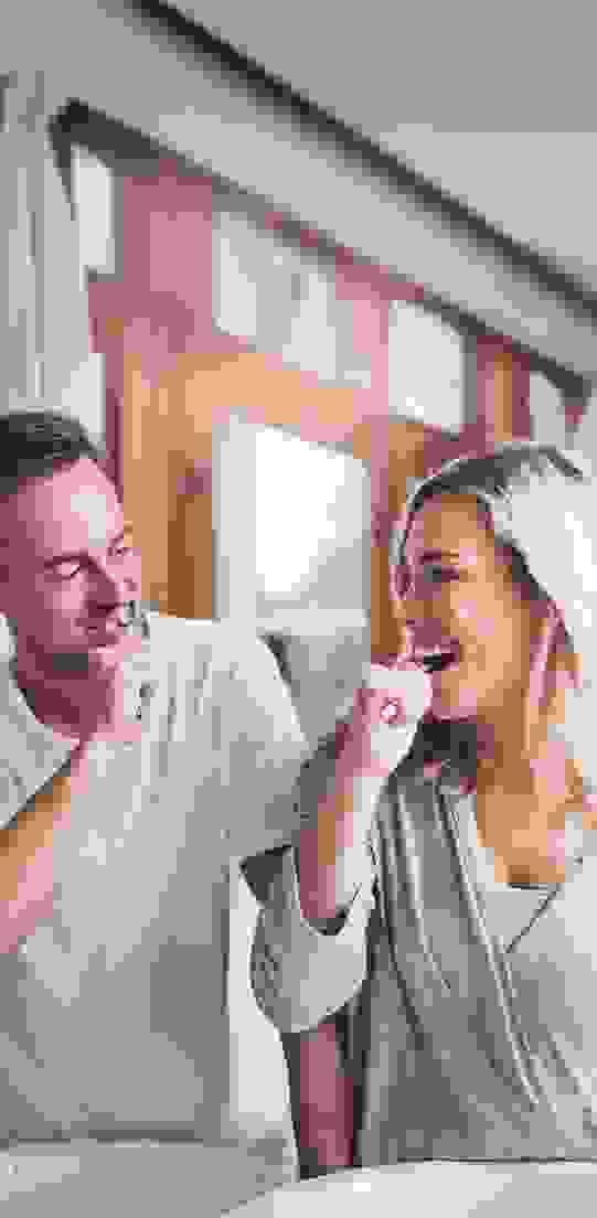 Due persone si lavano i denti fianco a fianco. Sorridono e sembrano divertirsi. La luce del giorno entra dalle finestre con tende bianche.