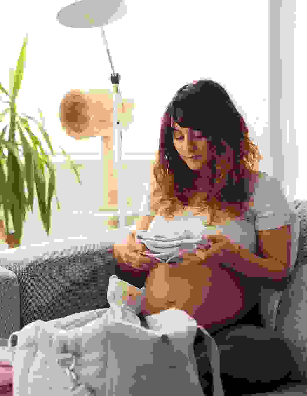 Eine Schwangere sitzt auf dem Sofa und packt sorgfältig die Spitaltasche fürs Baby – ein Schritt näher zur Geburt.
