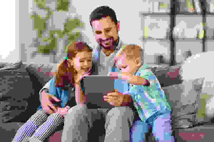 Ein Vater sitzt mit seinen zwei Kindern auf dem Sofa und schaut auf ein Tablet – ein Symbol für Nähe, Unterstützung und informierte Entscheidungen.