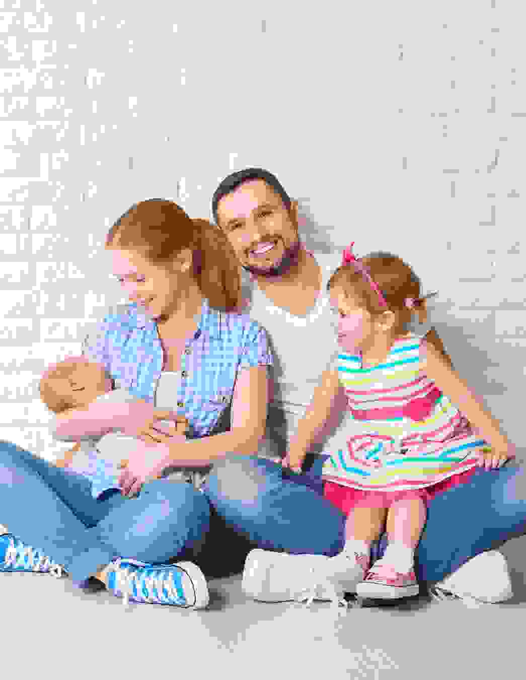 Una famiglia di quattro persone (padre, madre, bambina piccola e neonato) è seduta a gambe incrociate sul pavimento. L’immagine rappresenta la sicurezza e la coesione familiare offerte dalla soluzione cassa malati PRO LIFE.