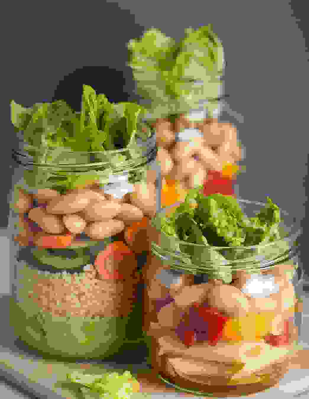 Drei Gläser gefüllt mit geschichtetem Salat aus Couscous, Pasta, Gemüse, Bohnen, Oliven und Blattsalat – ideal für unterwegs oder als Vorrat.
