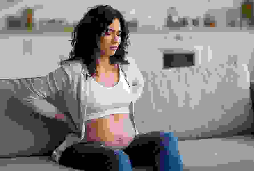 Eine Schwangere sitzt auf dem Sofa und hält sich schmerzhaft den Rücken – typische Beschwerden im letzten Trimester.