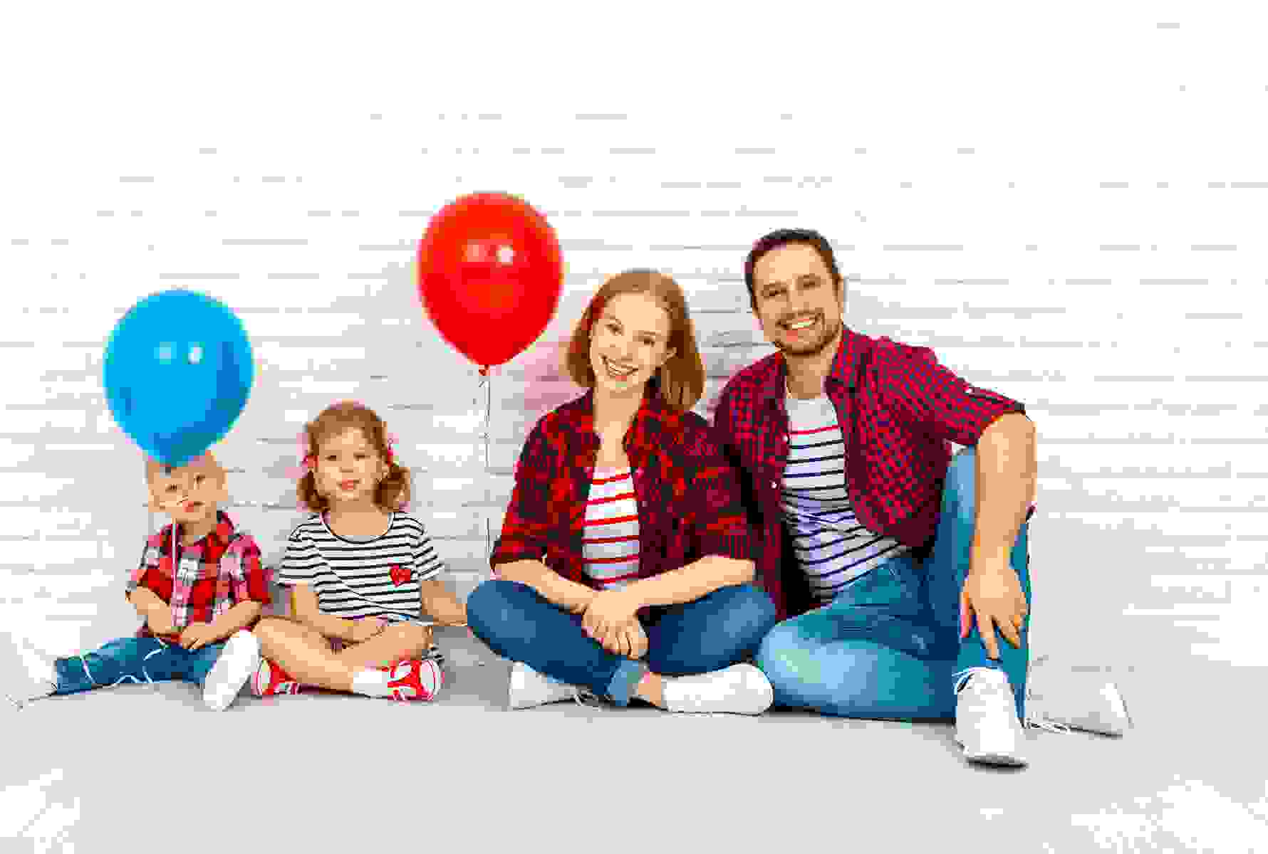 La même famille est assise détendue devant un mur en briques blanches, les enfants tiennent un ballon rouge et un ballon bleu – un moment plein de légèreté.