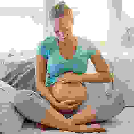 Eine strahlende Schwangere sitzt im Schneidersitz auf dem Sofa und hält liebevoll ihren Bauch – ein Moment der Vorfreude und inneren Ruhe kurz vor dem grossen Ereignis.