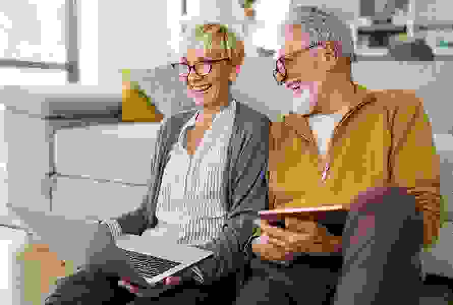 Un couple âgé et souriant est assis sur le canapé. La femme regarde son ordinateur portable – prête à s’informer et à plonger dans l’univers de PRO LIFE.