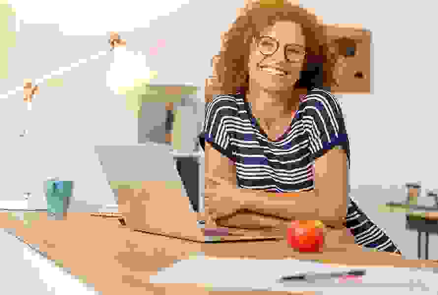 Eine Person sitzt an einem Schreibtisch mit Laptop und lächelt in die Kamera. Auf dem Tisch liegen ein Apfel, eine Lampe und Papiere. Im Hintergrund sind Bücherregale und Notizen an der Wand zu sehen – ein typisches Homeoffice-Setting.