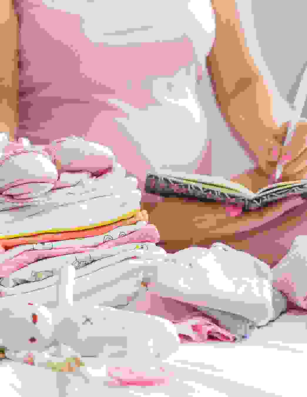 Eine schwangere Frau macht Notizen in ein Buch, im Vordergrund liegt liebevoll gefaltete Babykleidung – Erinnerungen für die Zukunft entstehen.