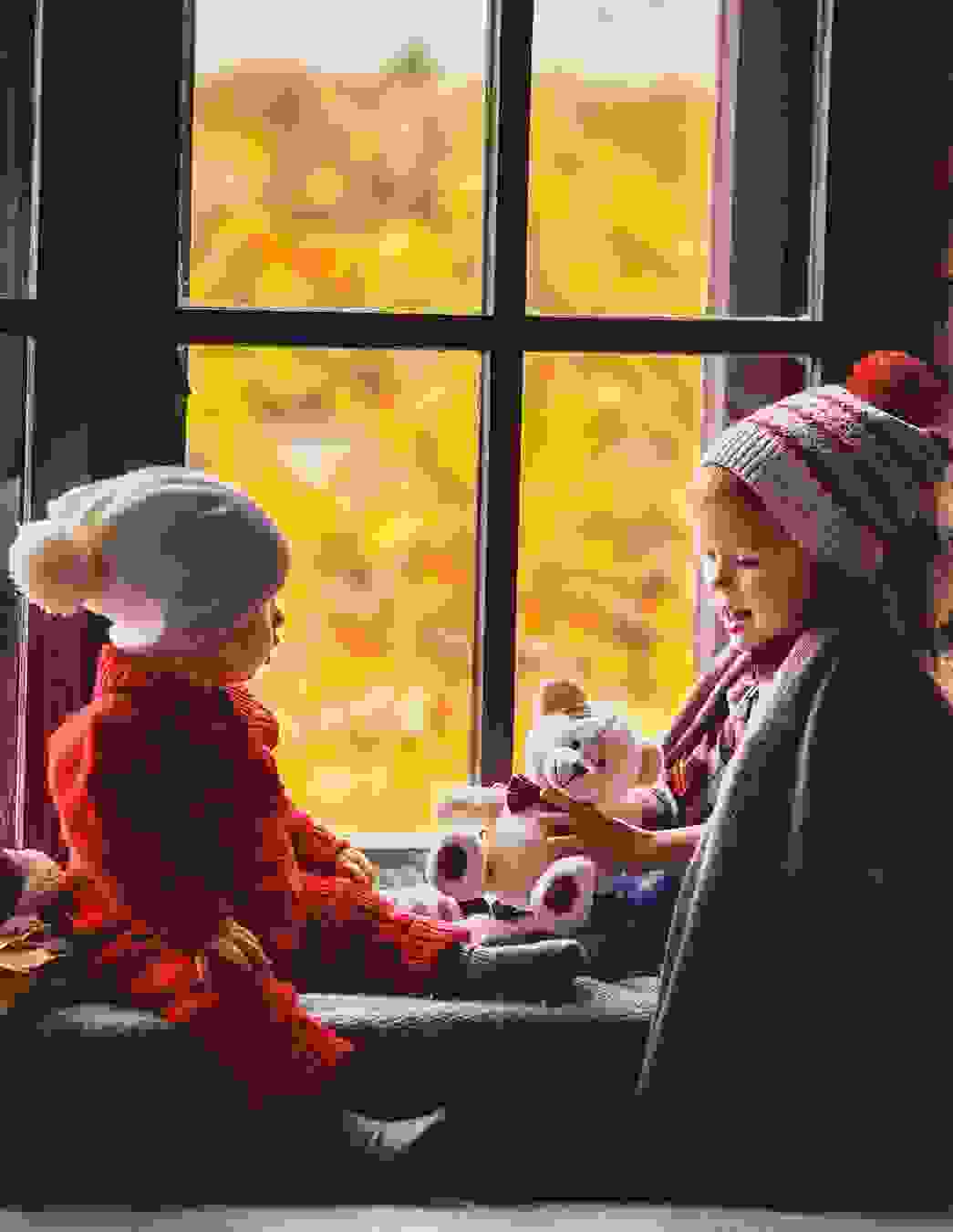 Ein Mädchen und ein Junge in Winterkleidung sitzen gemütlich auf der Fensterbank, das Mädchen hält einen Teddybären – ein Moment voller Wärme und Geborgenheit.