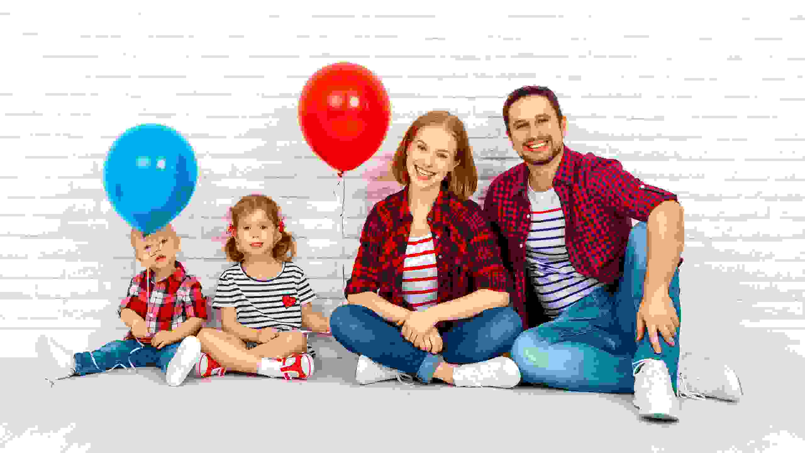 Eine vierköpfige Familie sitzt entspannt vor einer hellen Backsteinwand. Die Kinder halten bunte Ballone – ein Symbol für Leichtigkeit und Lebensfreude mit PRO LIFE.