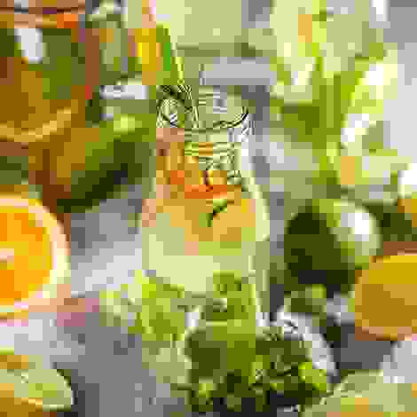 Flaschen mit Infused Water umgeben von frischen Zutaten wie Rosmarin, Melisse, Orangen, Zitronen und Limetten – lecker, kalorienarm und durstlöschend.