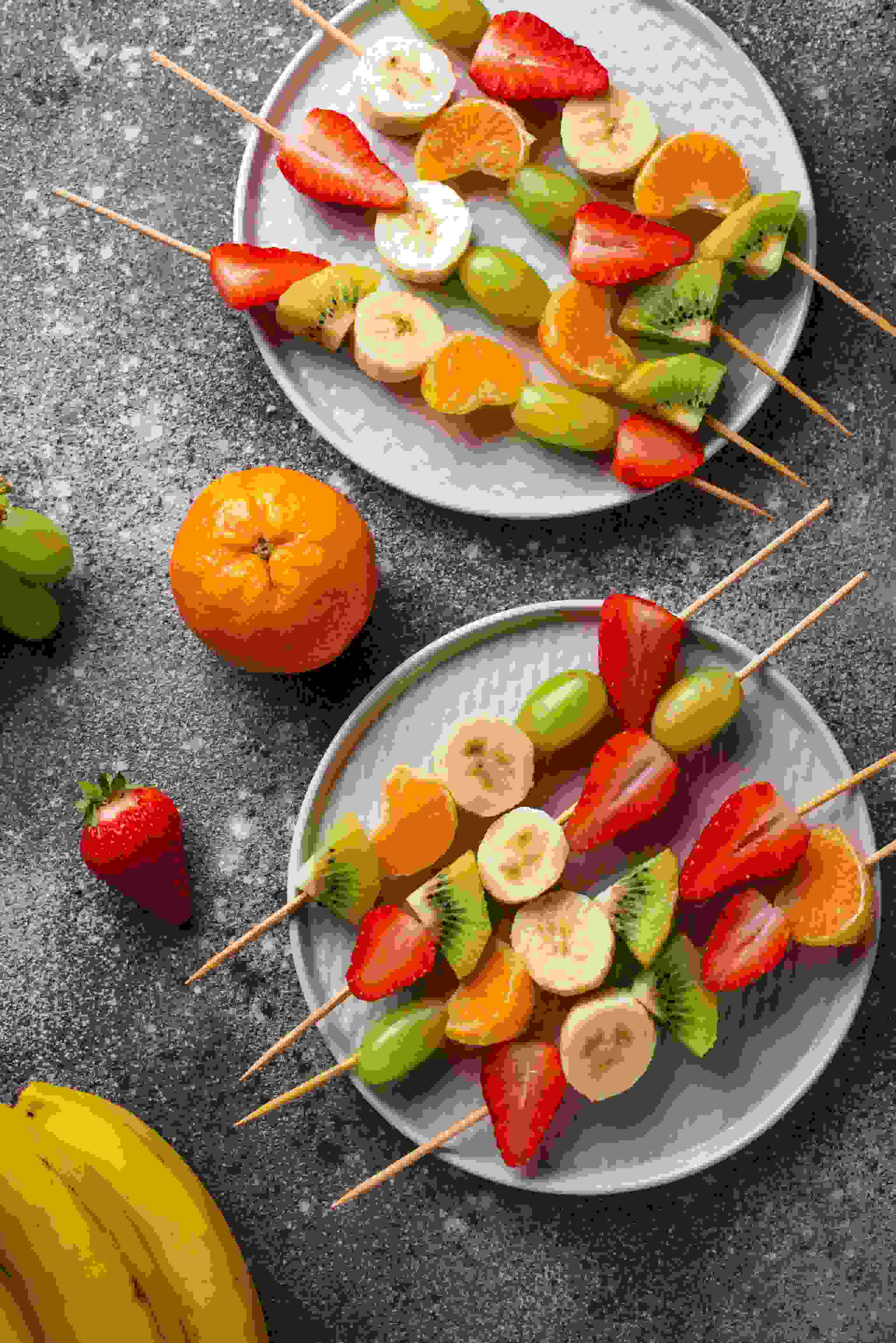 Zwei Teller mit bunten Fruchtspiessen aus Erdbeeren, Trauben, Mandarinen, Kiwi und Bananen. Gesund, erfrischend und ideal gegen kleine Gelüste.