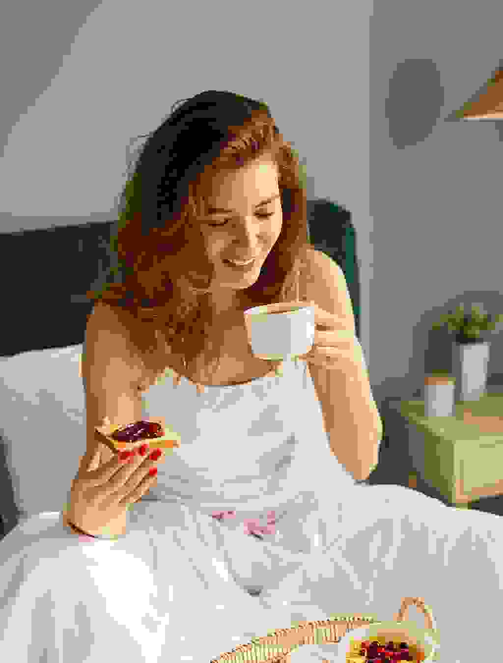 Eine Frau sitzt im Bett, hält eine Tasse in der einen und eine Scheibe Toast mit Konfitüre in der anderen Hand – ein Moment der Ruhe trotz Morgenübelkeit.