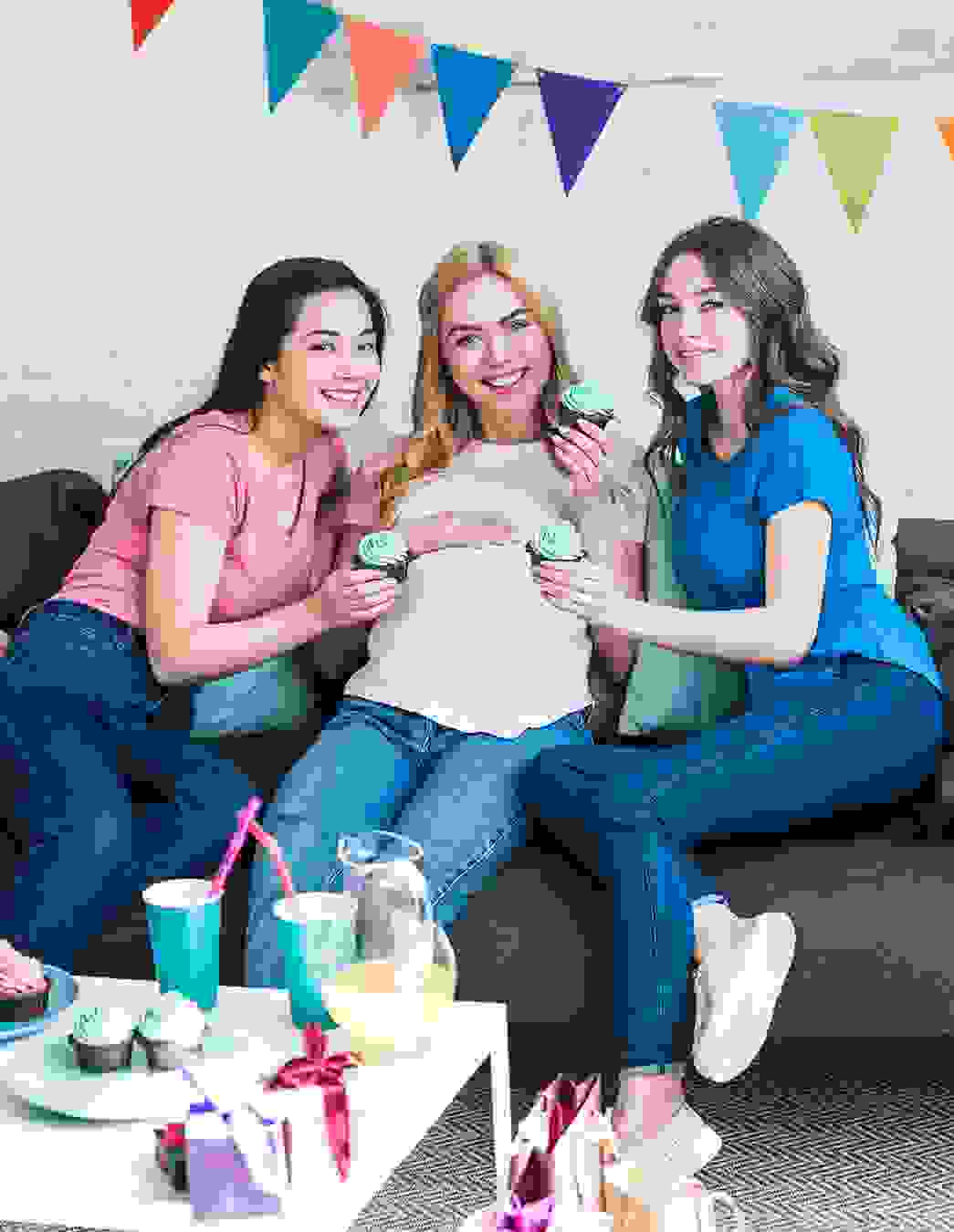 Eine Schwangere sitzt mit zwei Freundinnen auf dem Sofa – Babyparty-Stimmung mit Snacks, Gesprächen und kleinen Spielen.
