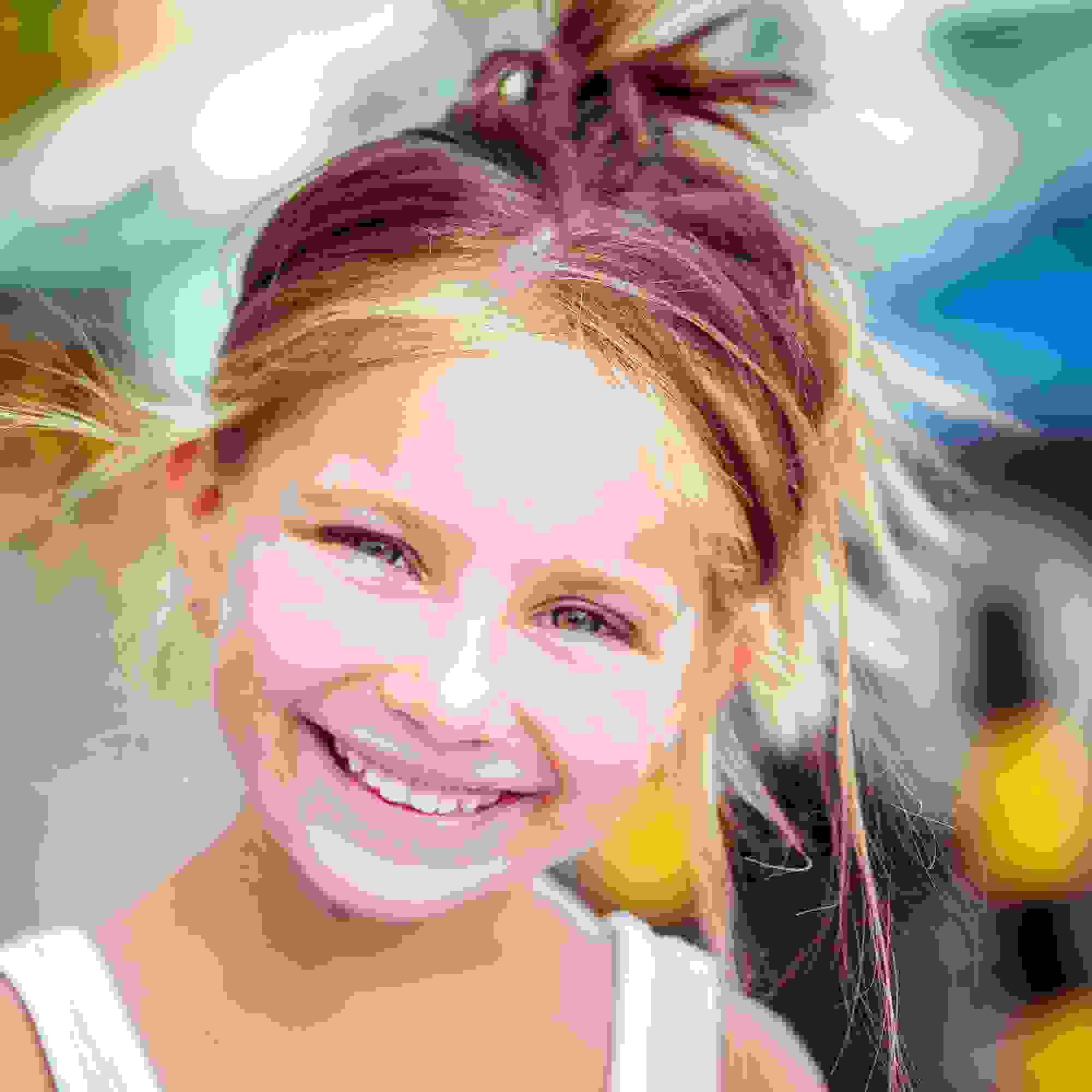 Ein blondes Kind mit hochgebundenen Haaren lächelt in die Kamera. Es trägt ein weisses Oberteil und befindet sich im Freien. Der Hintergrund ist bunt und unscharf.