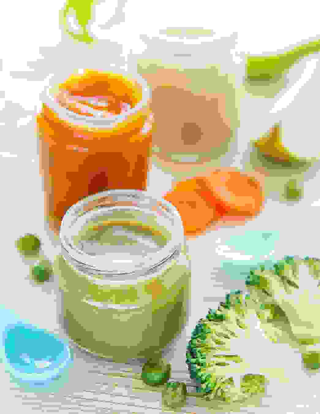 Tre vasetti di pappa fatta in casa sono disposti su un tavolo di legno chiaro, accanto a cucchiaini per bebè, broccoli, piselli, carote e una mela – tutto è pronto per le prime cucchiaiate di divertimento

