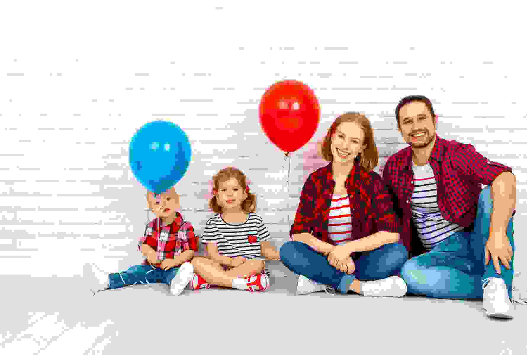 Die gleiche Familie sitzt entspannt vor einer weissen Backsteinmauer, die Kinder halten einen roten und blauen Ballon – ein Moment voller Leichtigkeit.