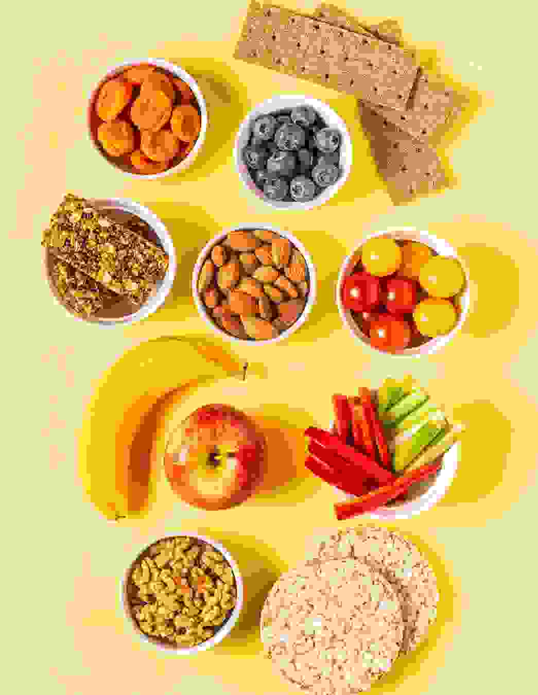 Auf dem Bild sehen Sie bunte Schälchen mit Aprikosen, Blaubeeren, Müesliriegeln, Mandeln, Cherrytomaten und Gemüsesticks – alles zusammen mit Vollkornknäckebrot, Reiswaffeln und frischem Obst. Perfekt für kleine Gelüste zwischendurch!