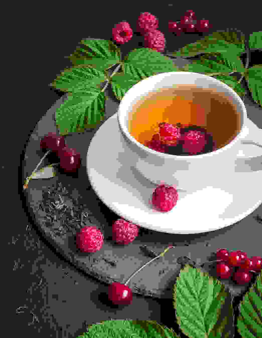 Eine Tasse Himbeerblättertee mit zwei frischen Himbeeren, serviert auf einer Schieferplatte. Daneben liegen frische sowie getrocknete Himbeerblätter und Himbeeren.