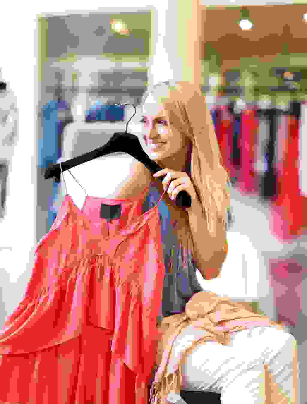 Blonde Frau hält ein rotes Trägerkleid hoch, zeigt die Freude über bequeme und stylische Umstandsmode in der 14. Schwangerschaftswoche.