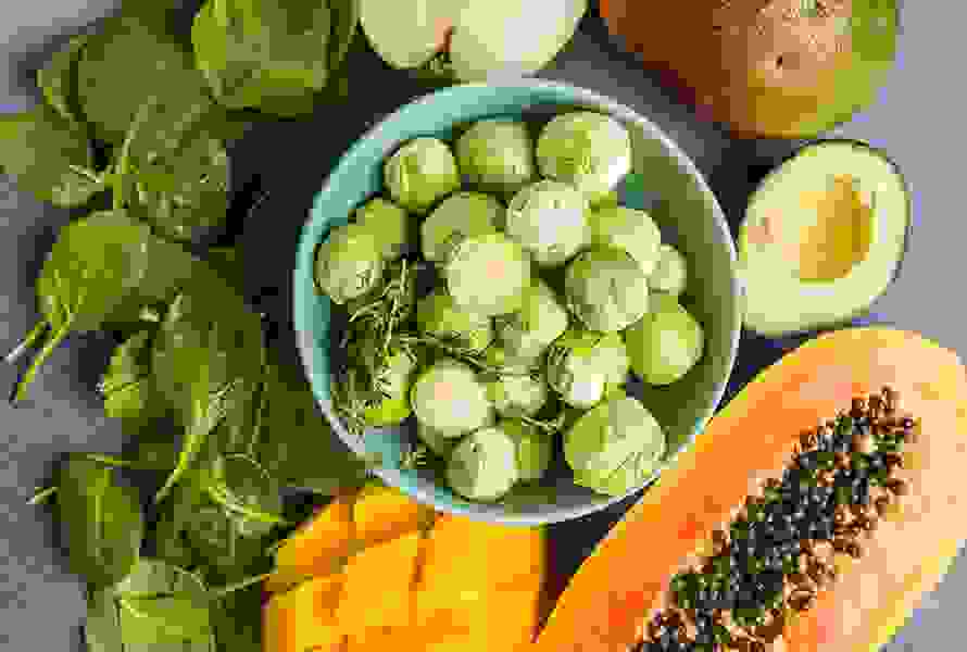 Eine Auswahl an folatreichen Lebensmitteln: Rosenkohl, Papaya, Mango, Spinat und Apfel – ideale Begleiter in der Schwangerschaft.