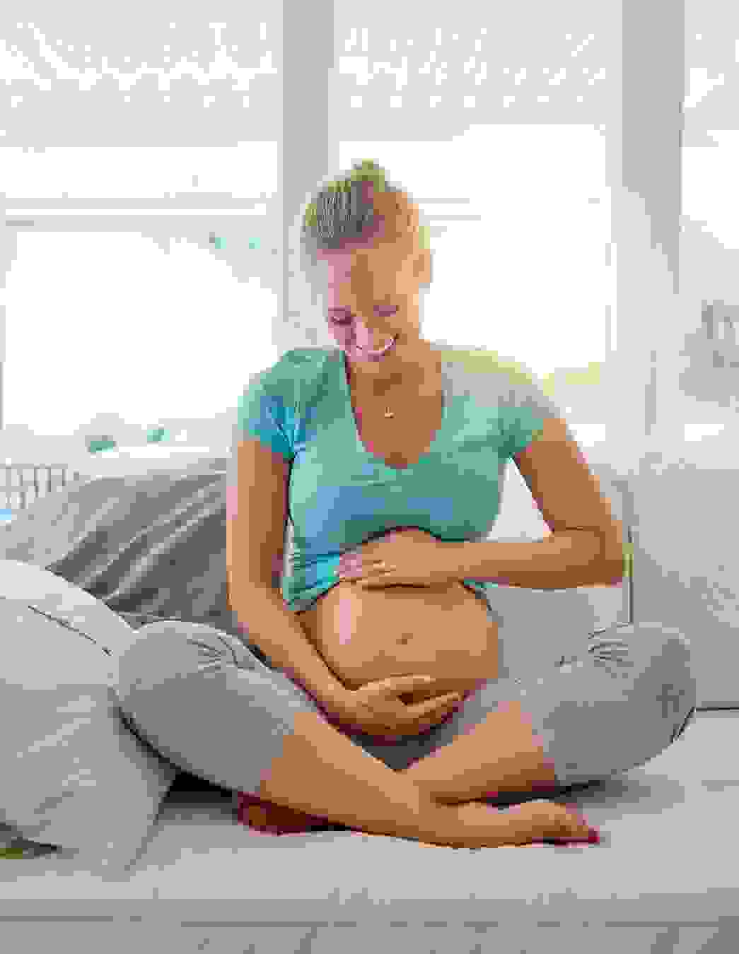 Eine glücklich lächelnde schwangere Frau sitzt im Schneidersitz auf dem Sofa und hält liebevoll ihren Bauch. Dieser Moment steht für die Unterstützung durch Krankenkassenleistungen während der Schwangerschaft in der Schweiz.
