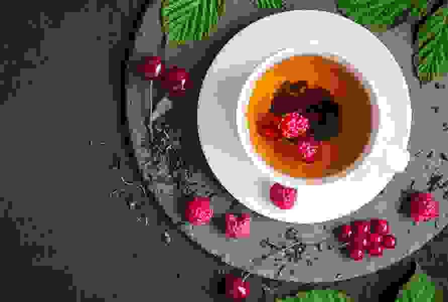 Eine Tasse Himbeerblättertee, dekorativ umgeben von frischen und getrockneten Himbeerblättern sowie Himbeeren – diese Woche ist eine Einnahmepause angesagt.