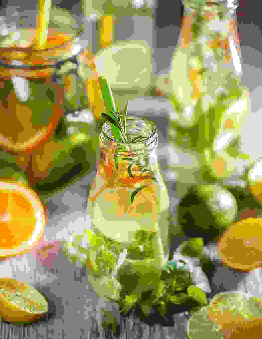 Flaschen mit Infused Water umgeben von frischen Zutaten wie Rosmarin, Melisse, Orangen, Zitronen und Limetten – lecker, kalorienarm und durstlöschend.