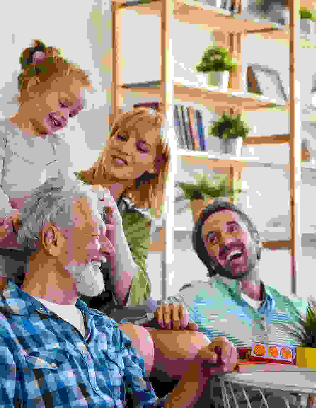 In einem warmen, wohnlichen Raum mit Bücherregalen und Pflanzen teilen vier Menschen einen fröhlichen Moment. Ein älterer Mann sitzt im Vordergrund, während eine Frau und ein Kind liebevoll mit ihm interagieren. Eine weitere erwachsene Person beobachtet die Szene lächelnd. Ein Bild voller Nähe und Lebensfreude.