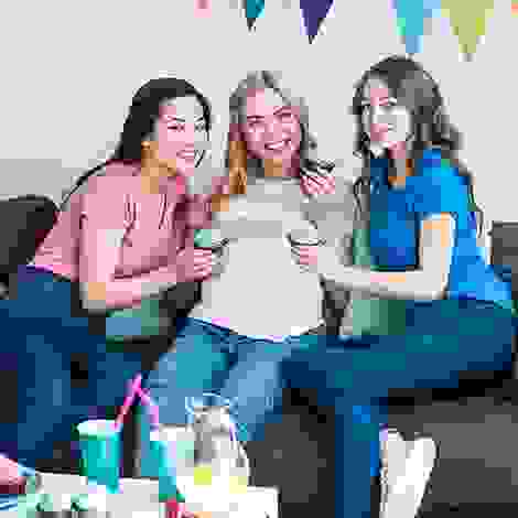 Eine Schwangere sitzt mit zwei Freundinnen auf dem Sofa – Babyparty-Stimmung mit Snacks, Gesprächen und kleinen Spielen.
