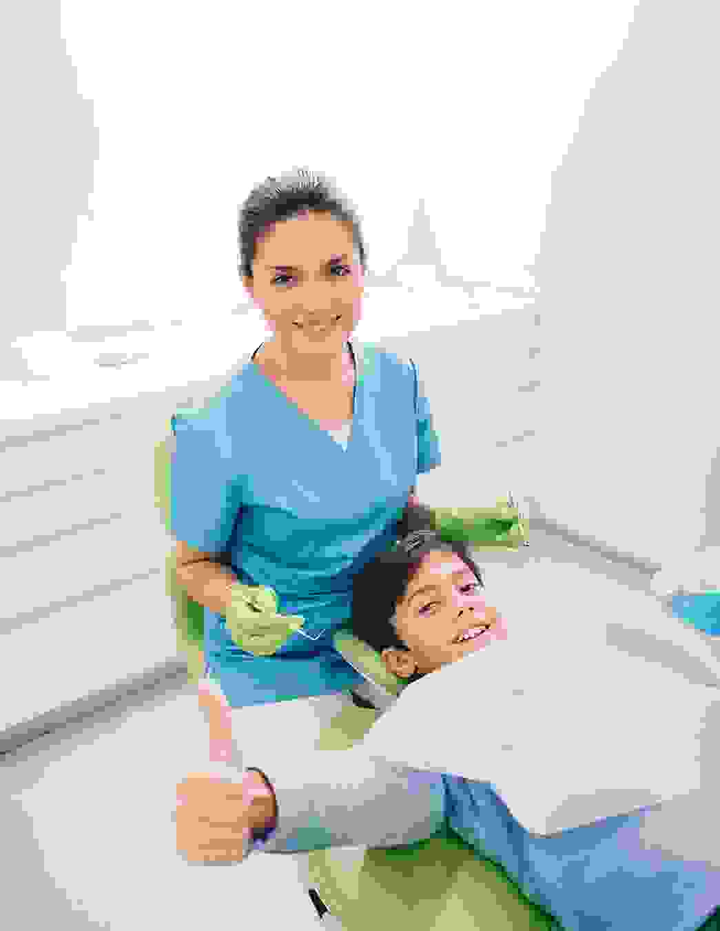 Une jeune dentiste soigne un patient, tandis qu’un garçon sourit à la caméra en levant le pouce – un signe fort de confiance et de bons soins.