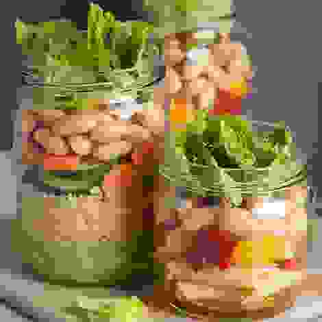 Drei Gläser gefüllt mit geschichtetem Salat aus Couscous, Pasta, Gemüse, Bohnen, Oliven und Blattsalat – ideal für unterwegs oder als Vorrat.