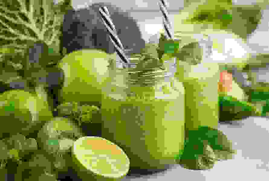 Due bicchieri di frullato verde circondati da broccoli, mela verde, kiwi e lime – rinfrescante, ricco di fibre e ideale contro gli attacchi di fame.

