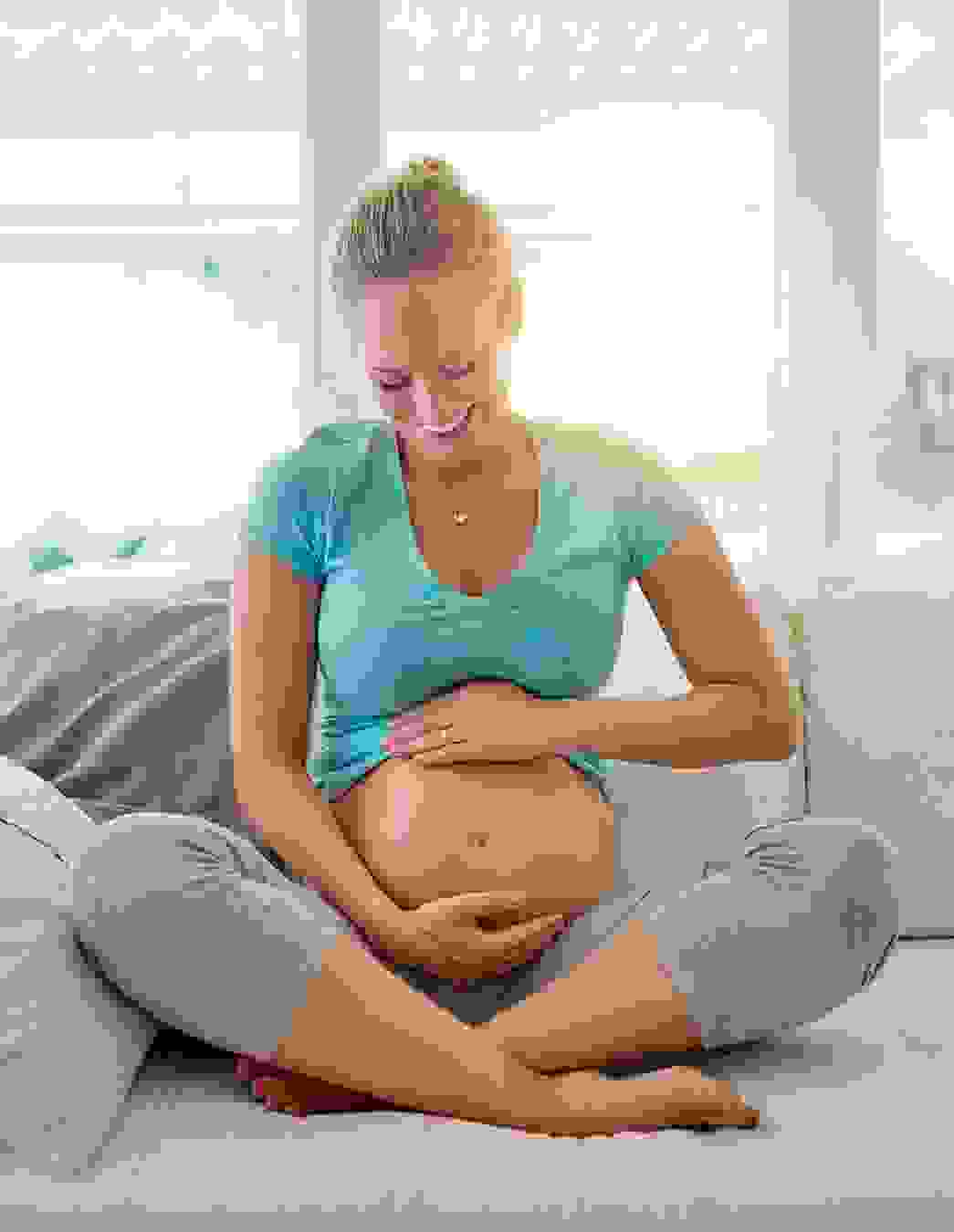 Eine strahlende Schwangere sitzt im Schneidersitz auf dem Sofa und hält liebevoll ihren Bauch – ein Moment der Vorfreude und inneren Ruhe kurz vor dem grossen Ereignis.