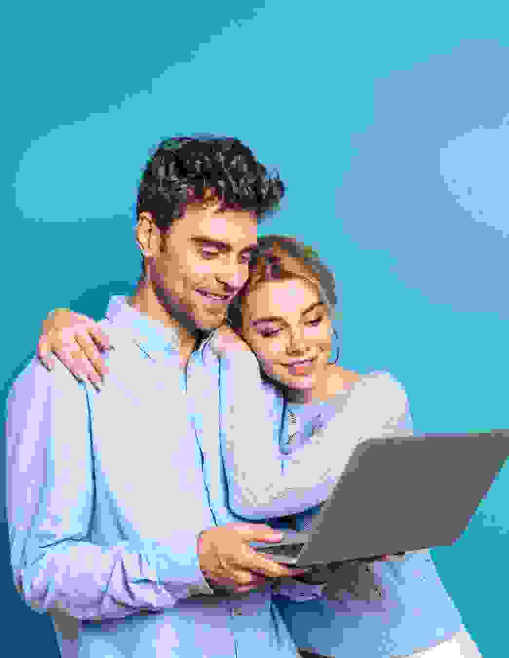 L’immagine mostra due persone che guardano insieme un computer portatile. Entrambe indossano abiti azzurri e si trovano davanti a uno sfondo blu. Una persona tiene il computer portatile, mentre l’altra le cinge le spalle con il braccio.