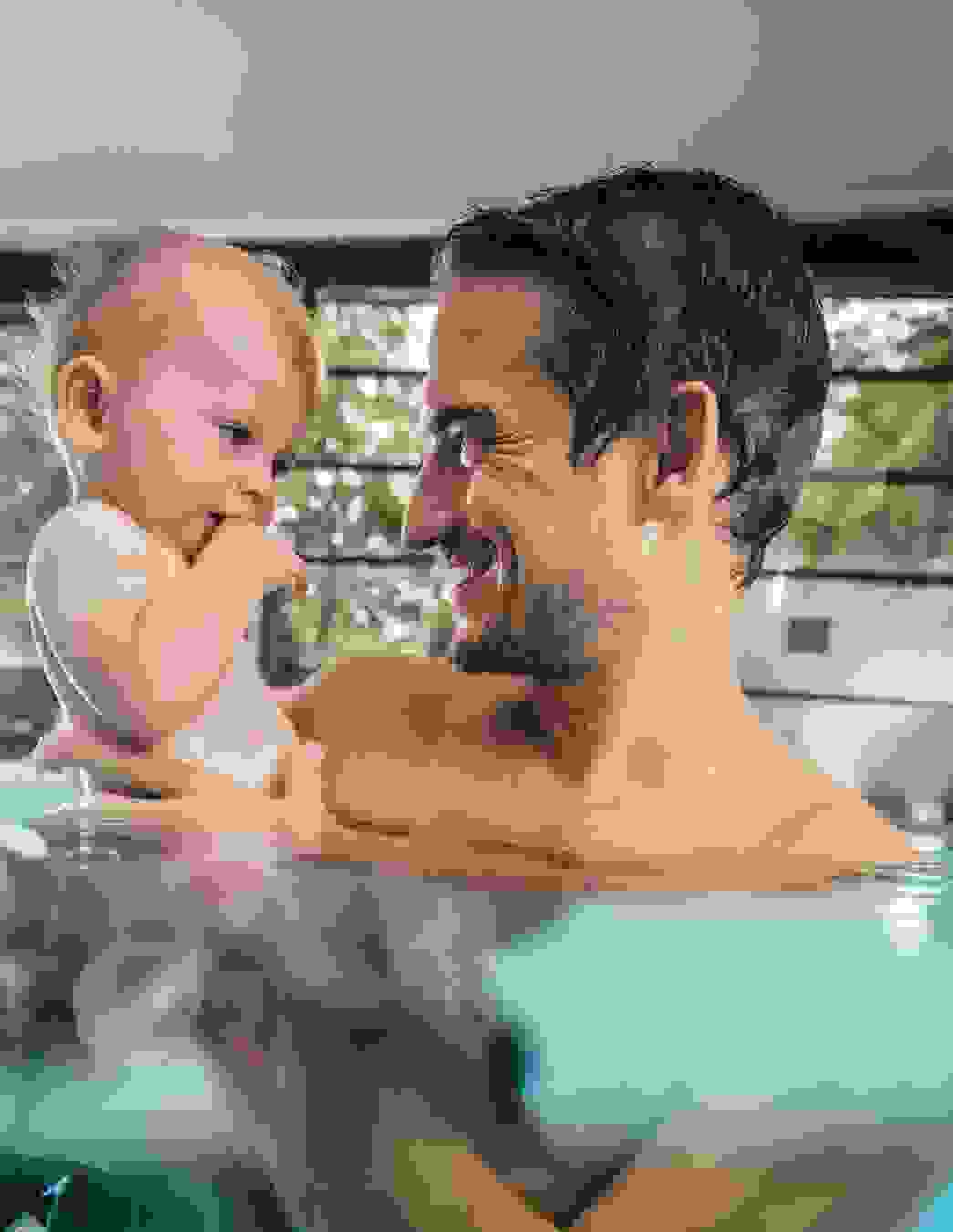 Un uomo tiene in braccio il suo bambino piccolo, entrambi ridono nell’acqua calda di una piscina coperta – vicinanza e fiducia grazie al nuoto per bebè.


