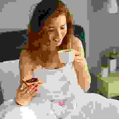 Eine Frau sitzt im Bett, hält eine Tasse in der einen und eine Scheibe Toast mit Konfitüre in der anderen Hand – ein Moment der Ruhe trotz Morgenübelkeit.