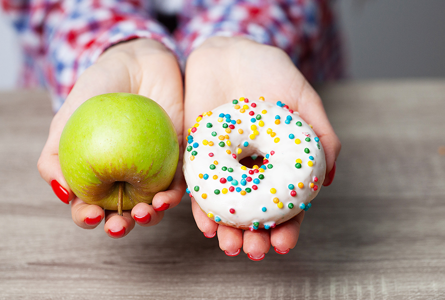 Entscheidung zwischen gesund und süss Eine Frau hält einen grünen Apfel in der einen und einen Donut mit weisser Glasur und bunter Deko in der anderen Hand – eine symbolische Darstellung der bewussten Ernährung in der Schwangerschaft.
