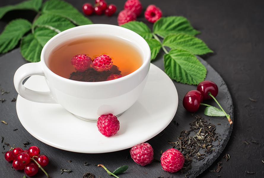 Himbeerblättertee richtig geniessen Eine dampfende Tasse Himbeerblättertee, dekorativ umgeben von frischen Himbeeren und Blättern – bitter-herb im Geschmack, aber hilfreich zur Geburtsvorbereitung.