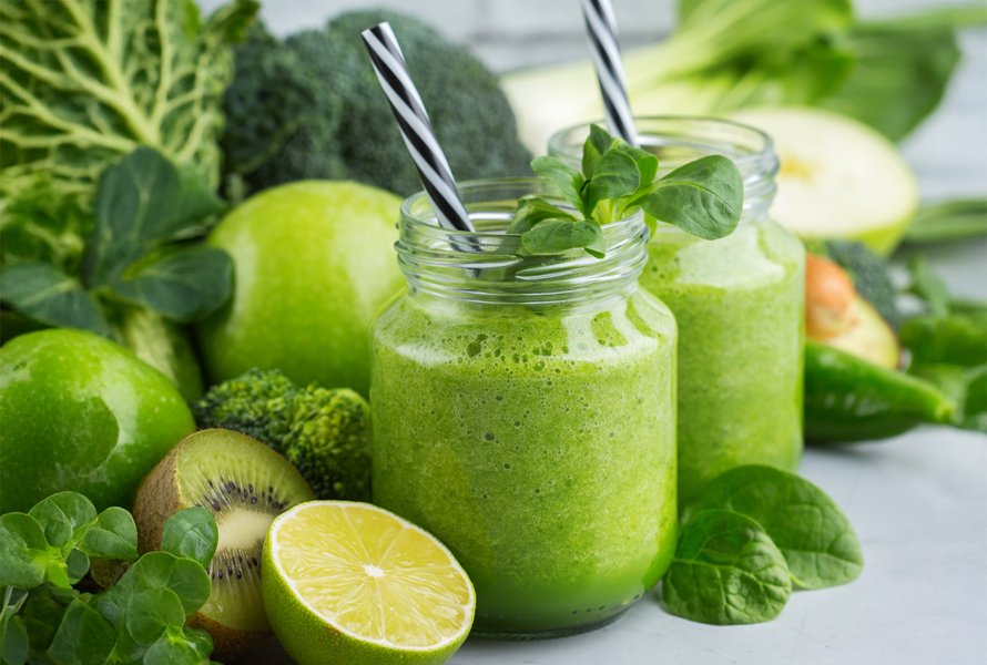 Due bicchieri di frullato verde circondati da broccoli, mela verde, kiwi e lime – rinfrescante, ricco di fibre e ideale contro gli attacchi di fame.

