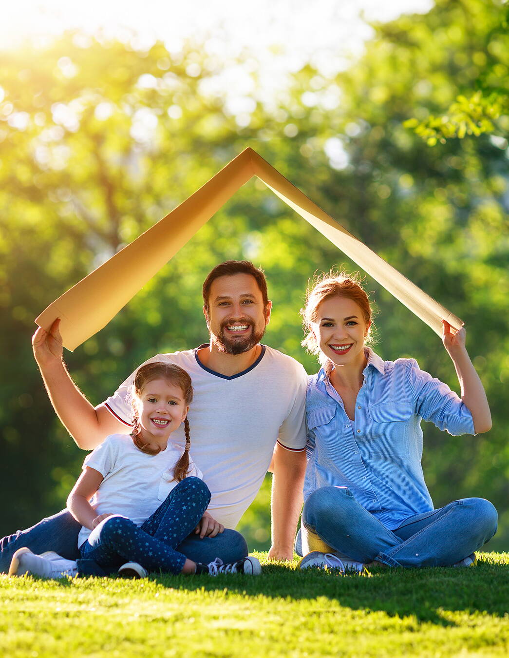 Un couple est assis avec leur petite fille sur une pelouse verte et tient au-dessus de leurs têtes un morceau de carton en forme de toit – un symbole de sécurité et de protection.