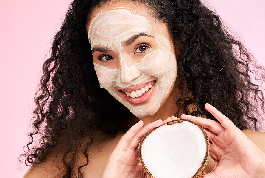 DIY-Gesichtsmaske für empfindliche Haut Junge Frau mit selbst aufgetragener Gesichtsmaske hält eine halbierte Kokosnuss – natürliche Zutaten für strahlende Haut und Wohlgefühl.
