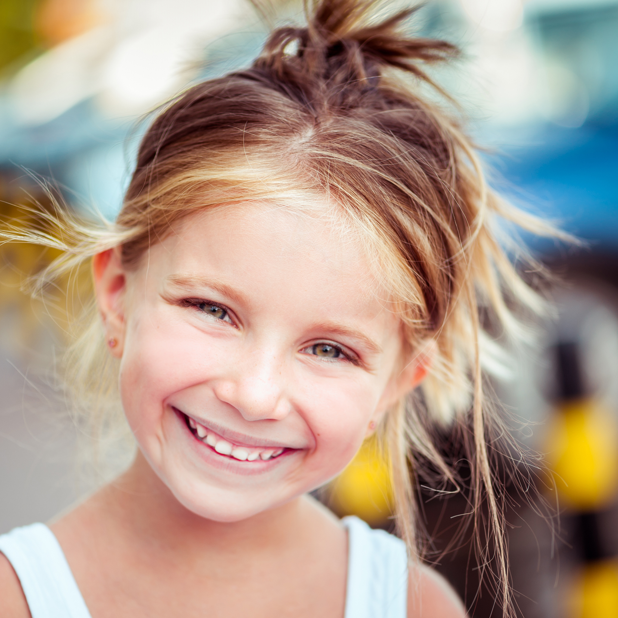 Ein blondes Kind mit hochgebundenen Haaren lächelt in die Kamera. Es trägt ein weisses Oberteil und befindet sich im Freien. Der Hintergrund ist bunt und unscharf.